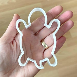 May include: White plastic turtle cookie cutter. The cutter is shaped like a turtle with a rounded shell and four legs.