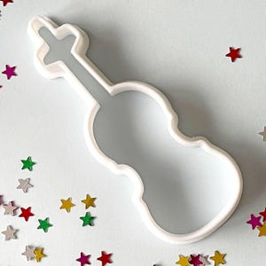 Pode incluir: Cortador de biscoitos de plástico branco em forma de violino. O cortador está sobre um fundo azul claro com confeitos em forma de estrela coloridos espalhados.
