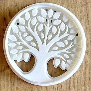 May include: A white, circular cookie cutter featuring a detailed tree design. The tree has a trunk and branches filled with leaf shapes, all within a round frame. The cutter is made of a solid, opaque white material.