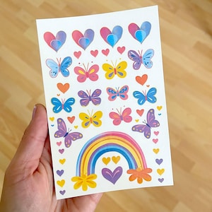 Op de afbeelding: Een vel kleurrijke stickers met vlinders, harten en een regenboog. De stickers zijn in verschillende kleuren, waaronder roze, blauw, geel en paars. De regenboog heeft gele, oranje, rode, paarse en blauwe strepen.