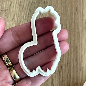 May include: A white plastic cookie cutter in the shape of a llama. The cutter is held in a hand, with a wooden surface in the background. The llama shape has a head, body, and legs.
