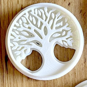 Puede incluir: Un cortador de galletas circular blanco con un diseño detallado del árbol de la vida. Las ramas y hojas del árbol están intrincadamente talladas dentro de un marco circular. El cortador está hecho de un material blanco opaco.