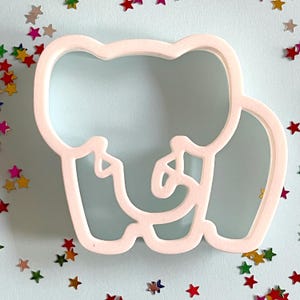May include: White plastic cookie cutter shaped like an elephant. The elephant has a trunk, ears, and legs. The cookie cutter is on a light blue background.