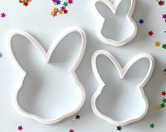 May include: Three white plastic bunny cookie cutters of different sizes on a light blue background with colorful star-shaped sprinkles.