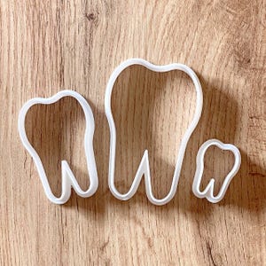 Puede incluir: Tres cortadores de galletas blancos con forma de diente de diferentes tamaños están dispuestos sobre una superficie de madera. Los cortadores tienen forma de molares e incisivos, perfectos para proyectos de repostería con temática dental.