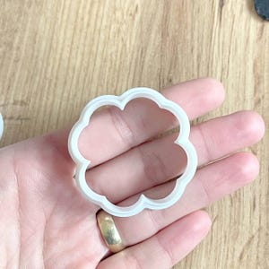 Può includere: Tagliapasta bianco a forma di nuvola con bordo smerlato. Il tagliapasta è tenuto in mano. Lo sfondo è una superficie di legno marrone chiaro.