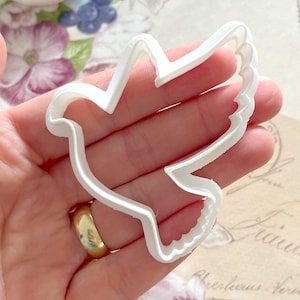 May include: White plastic dove cookie cutter. The cutter has a detailed design with a textured edge.