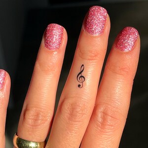 Op de afbeelding: Een close-up van een vinger met roze glitternagellak en een kleine zwarte viool-sleutel tattoo.