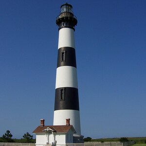 K&ouml;nnte beinhalten: Ein hoher, konischer Leuchtturm mit abwechselnden horizontalen schwarzen und wei&szlig;en Streifen. Das Bauwerk ist mit einer gl&auml;sernen Laterne gekr&ouml;nt und verf&uuml;gt an der Basis &uuml;ber ein kleines, wei&szlig;es einst&ouml;ckiges Geb&auml;ude mit braunem Dach.