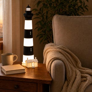 K&ouml;nnte beinhalten: Eine hohe, zylindrische Leuchtturm-Lampe mit abwechselnden horizontalen schwarzen und wei&szlig;en Streifen, gekr&ouml;nt von einer schwarzen Laterne. Ein kleines, wei&szlig;es, hausf&ouml;rmiges Nachtlicht steht an der Basis, beide strahlen ein warmes Licht aus.