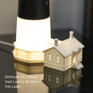 K&ouml;nnte beinhalten: Ein wei&szlig;es Miniaturhaus mit Schornstein und Fenstern, das leuchtet und neben einer hohen Leuchtturmlampe steht. Der Text lautet: Detailed Oil House that lights up with the lamp. Tabletop Landmarks steht unten rechts im Bild.