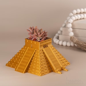 May include: A gold-colored, 3D printed planter shaped like the Mayan pyramid of Chichen Itza. The planter has a small succulent plant growing in the top.