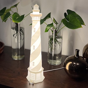 May include: A decorative lighthouse lamp with alternating beige and white stripes. The lamp has a detailed design, including windows and a top structure. The lamp is plugged in and illuminated, casting a warm glow. The lamp is on a wooden surface.