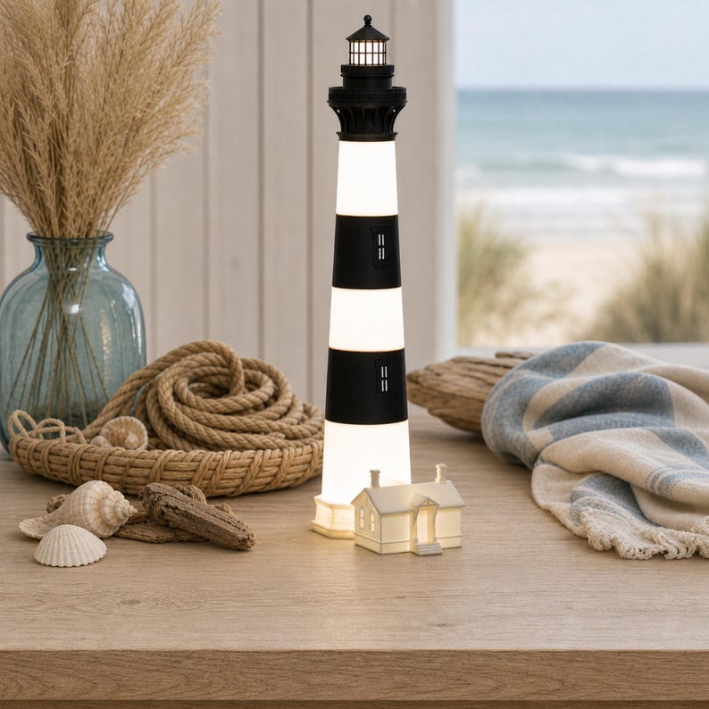 K&ouml;nnte beinhalten: Eine hohe, zylindrische Leuchtturm-Lampe mit abwechselnden horizontalen schwarzen und wei&szlig;en Streifen, gekr&ouml;nt von einer schwarzen Laterne, die ein sanftes Licht ausstrahlt. Daneben steht ein kleines, wei&szlig;es, beleuchtetes Haus mit Spitzdach.