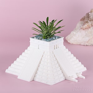 May include: White ceramic planter shaped like the Mayan pyramid of Chichen Itza, with a small green succulent plant growing inside. The planter has a textured surface and is decorated with small blue stones.