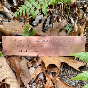 Może przedstawiać: Miedziana tabliczka z napisem "there is nowhere else to go there is no one else to be you are home." Tabliczka leży na brązowych liściach.