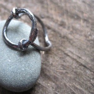 Può includere: Un anello d'argento con un designo a nodo unico. L'anello è in metallo e ha una finitura liscia e lucida.