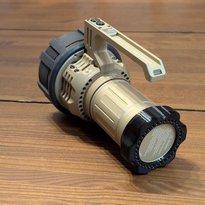 May include: A tan and black spotlight with a handle. The spotlight has a cylindrical body with a black bezel and a handle on top. The spotlight is designed for industrial or outdoor use.