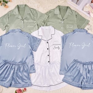 May include: Matching pajama sets in sage green, white, and blue. Each set includes a short-sleeved button-down shirt and shorts. The shirts are personalized with names like "Anna", "Sarah", and "Emily", and the blue sets say "Flower Girl".