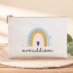 Op de afbeelding: Een witte canvas pouch met een gouden rits, met een kleurrijk regenboogontwerp met sterren en een hart. De naam "mrs addison" staat eronder. De pouch ligt op een geweven mat.