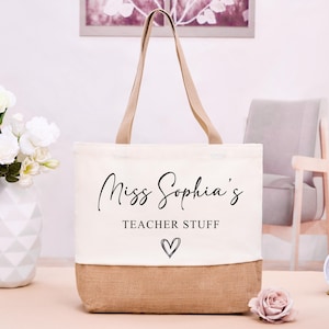 May include: Cream-colored tote bag with the text "Miss Sophia's TEACHER STUFF" and a heart graphic. The bottom of the bag is a natural burlap color, with tan handles. A vase of white flowers and a clock are in the background.