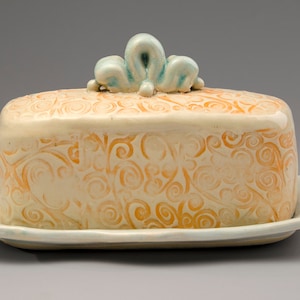 May include: Ceramic butter dish with a cream-colored base and lid, adorned with orange swirl patterns. The lid features a decorative, light blue accent. The dish sits on a matching, slightly scalloped tray, perfect for kitchen decor.