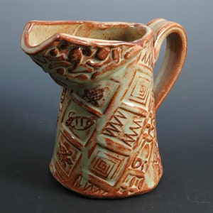 May include: A brown and green ceramic pitcher with a textured surface and a handle. The pitcher has a geometric pattern with a repeating design of squares and circles.