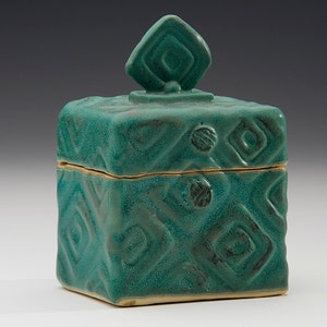May include: A teal ceramic box with a geometric pattern and a lid with a diamond-shaped knob.