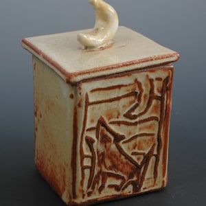 May include: A small, square ceramic box with a brown and white glaze. The box has a carved design of a stylized animal on the front and a crescent moon shaped lid.