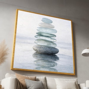 Puede incluir: Una obra de arte enmarcada que presenta una pila de piedras lisas, grises y azules. Las piedras están equilibradas unas sobre otras, reflejándose en el agua de abajo. La obra está encerrada en un marco dorado, creando una composición serena y equilibrada.