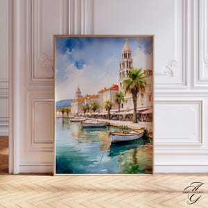 Może przedstawiać: Akwarela przedstawiająca nadmorskie miasteczko z łodziami zacumowanymi w turkusowej wodzie. Budynki z czerwonymi dachówkami i palmami wzdłuż brzegu pod błękitnym niebem. Grafika jest oprawiona w złotą ramę.