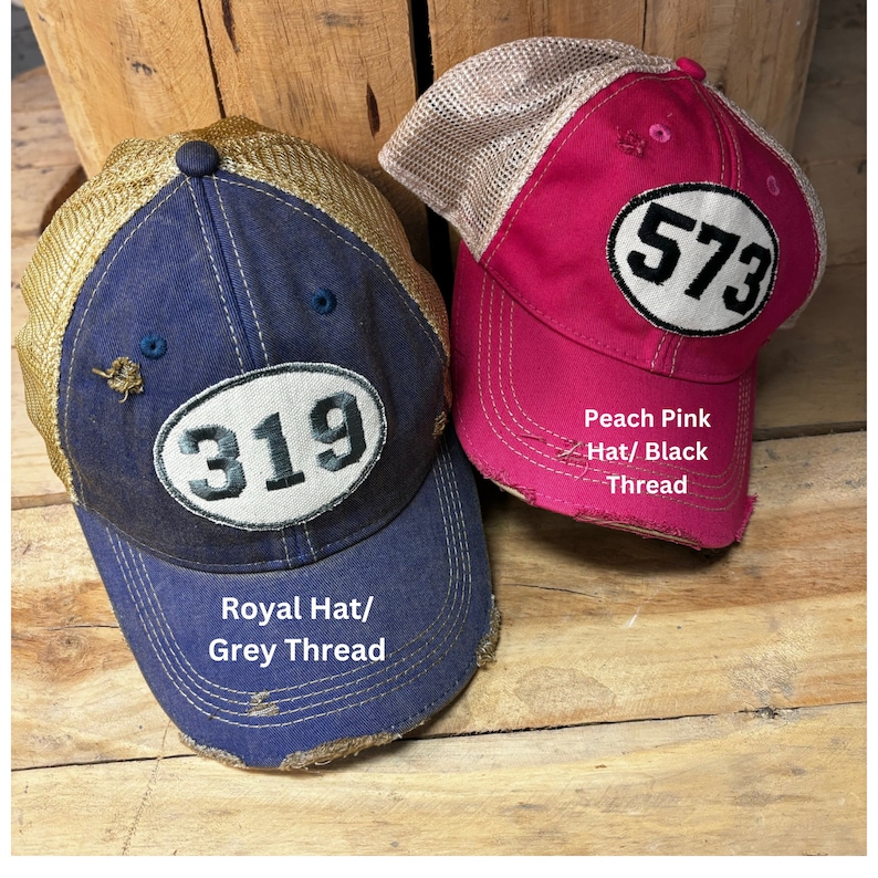 K&ouml;nnte beinhalten: Zwei Baseballkappen mit abgenutztem Stoff und einem Nummernaufn&auml;her auf der Vorderseite. Die Kappe links ist k&ouml;nigsblau mit einem goldenen Netzr&uuml;cken und einem wei&szlig;en Nummernaufn&auml;her mit der Nummer 319. Die Kappe rechts ist pfirsichfarben mit einem braunen Netzr&uuml;cken und einem wei&szlig;en Nummernaufn&auml;her mit der Nummer 573. Der Text auf den Kappen lautet "Royal Hat/Grey Thread" und "Peach Pink Hat/Black Thread".