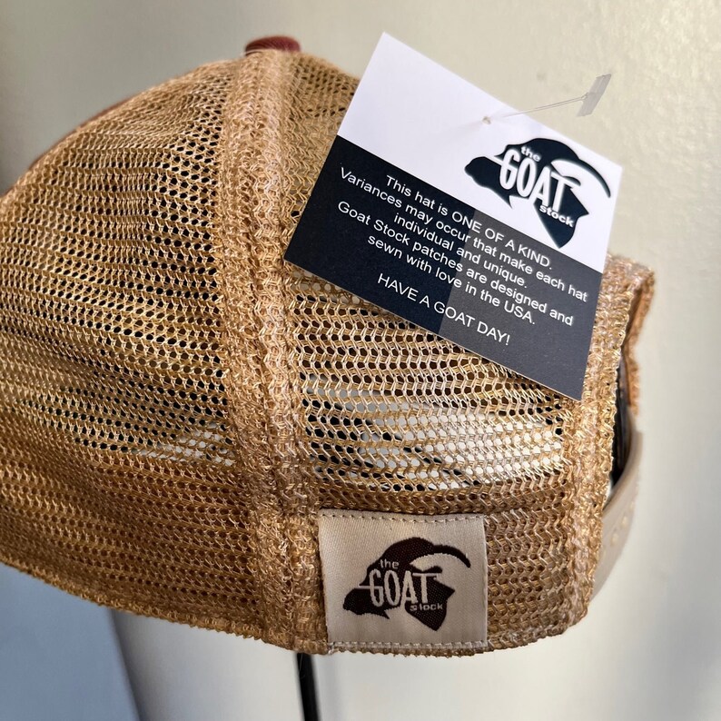 May include: A brown and tan mesh trucker hat with a brown leather strap and a black and white logo patch that says "the GOAT stock". The hat has a white label on the back with the same logo.