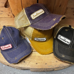 Op de afbeelding: Vier distressed trucker caps in blauw, goud en zwart, met mesh ruggen en gerafelde randen. Elke cap heeft een patch met een teamnaam: "Patriots", "Indians", "Tigers" en "Panthers".