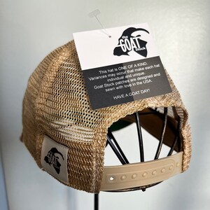K&ouml;nnte beinhalten: Eine braune und beige Mesh-Trucker-Kappe mit einem braunen Lederriemen und einem schwarzen Patch mit dem Text "the GOAT stock" und einer Ziegensilhouette. Die Kappe befindet sich auf einem Hutst&auml;nder. Dieser Hut ist EINZIGARTIG. Abweichungen k&ouml;nnen auftreten, die jeden Hut individuell und einzigartig machen. Goat Stock-Patches werden in den USA entworfen und mit Liebe gen&auml;ht. HABEN SIE EINEN SCH&Ouml;NEN TAG!