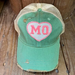 Op de afbeelding: Een groene en beige trucker-pet met een roze hartpatch waarop "MO" staat. De pet heeft een verweerde look en is gemaakt van een zacht, comfortabel materiaal.