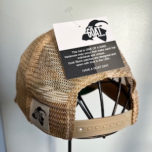 May include: A tan and brown mesh baseball cap with a black patch that says "The Goat Stock" and the text "This hat is ONE OF A KIND. Variances may occur that make each hat individual and unique. Goat Stock patches are designed and sewn with love in the USA. HAVE A GOAT DAY!"