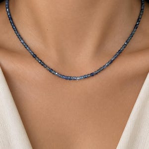 May include: A delicate choker necklace composed of small, faceted, deep blue beads. The beads exhibit a translucent quality with varying shades of navy and indigo, creating a subtle gradient effect around the neck. The necklace sits close to the base of the neck.