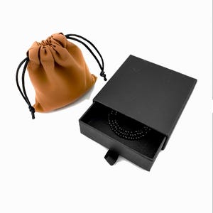 May include: A tan fabric drawstring pouch with black cords sits next to a black rectangular sliding jewelry box. The box is partially open, revealing a coiled black beaded necklace or bracelet resting inside the dark interior compartment.