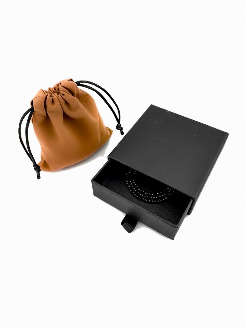 May include: A tan fabric drawstring pouch with black cords sits next to a black rectangular sliding jewelry box. The box is partially open, revealing a coiled black beaded necklace or bracelet resting inside the dark interior compartment.