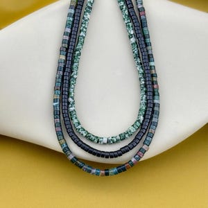 May include: A layered necklace set consisting of three strands of small cylindrical beads. One strand features mottled green and white stone beads, another contains solid black beads, and the third strand displays multicolored beads in shades of blue, green, and brown.
