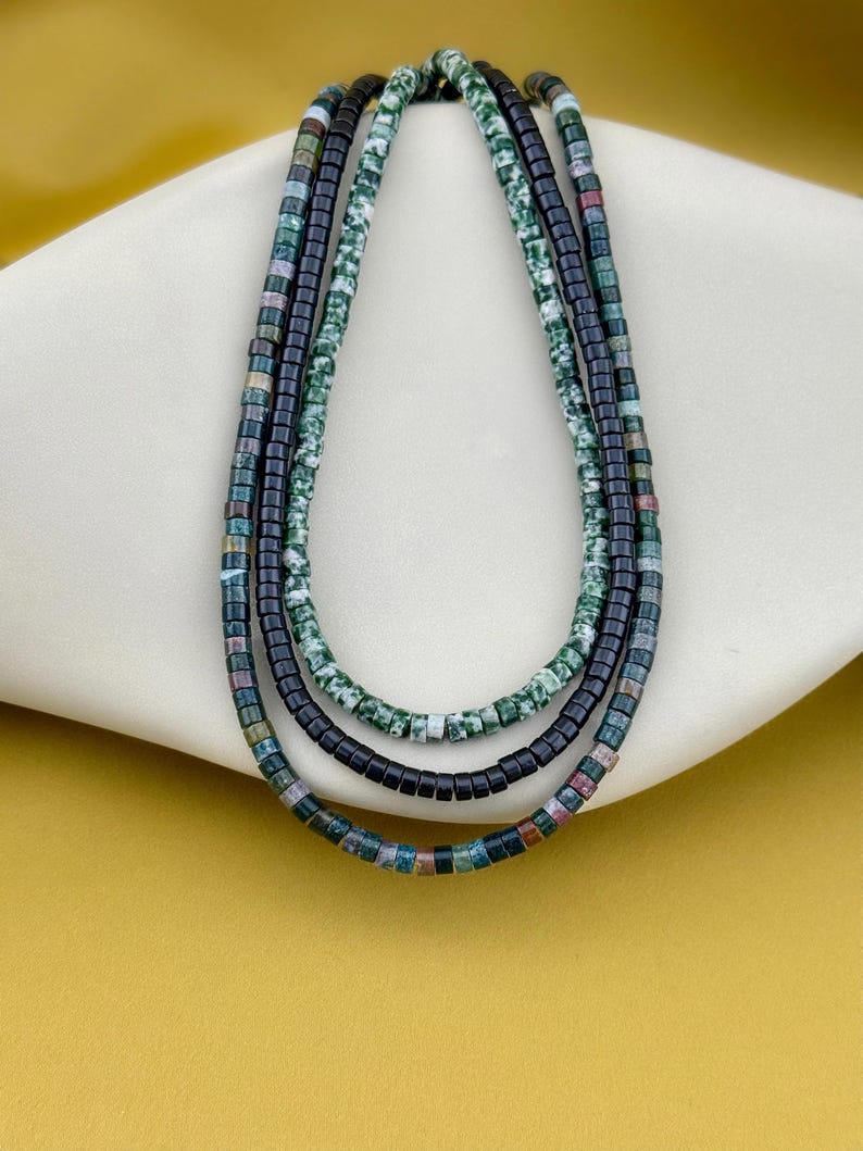 May include: A layered necklace set consisting of three strands of small cylindrical beads. One strand features mottled green and white stone beads, another contains solid black beads, and the third strand displays multicolored beads in shades of blue, green, and brown.