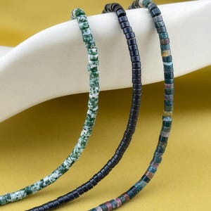 May include: Three strands of beaded jewelry featuring small cylindrical beads. One strand is mottled green and white, one is solid matte black, and one is a multicolored mix of green, brown, and grey earth-toned stone beads arranged in a repeating pattern.