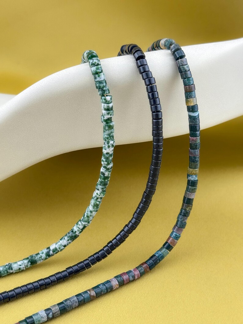 May include: Three strands of beaded jewelry featuring small cylindrical beads. One strand is mottled green and white, one is solid matte black, and one is a multicolored mix of green, brown, and grey earth-toned stone beads arranged in a repeating pattern.