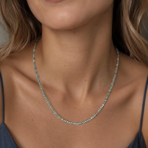 Puede incluir: Un delicado collar compuesto por pequeñas cuentas de color azul claro. El collar se lleva alrededor del cuello. El fondo es de un tono suave y neutro, que resalta la joya. El collar parece estar hecho de piedras preciosas. Mide unos 40 cm.