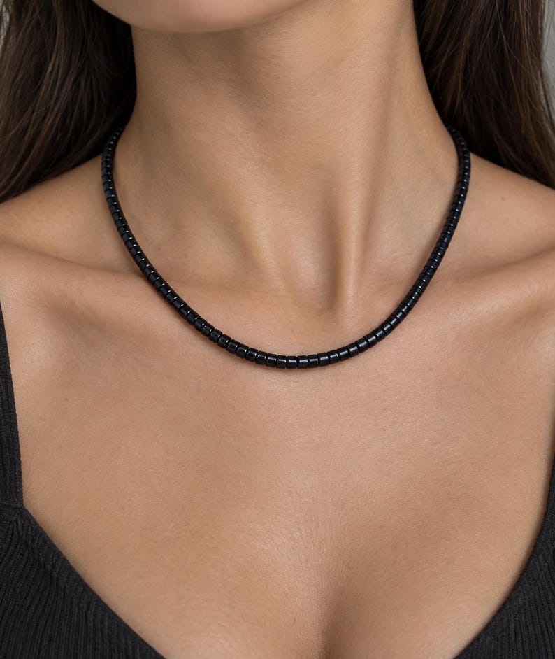 May include: A necklace composed of small, uniform, cylindrical black beads strung together in a single strand. The beads have a smooth, glossy finish and are arranged closely to form a continuous, circular chain that rests around the neck.