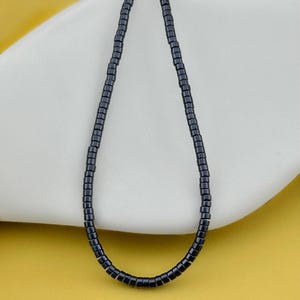 May include: A single strand necklace composed of small, cylindrical, dark charcoal-colored beads. The beads are uniform in shape and size, strung together to form a continuous loop that hangs in a simple, elongated oval shape against a smooth surface.