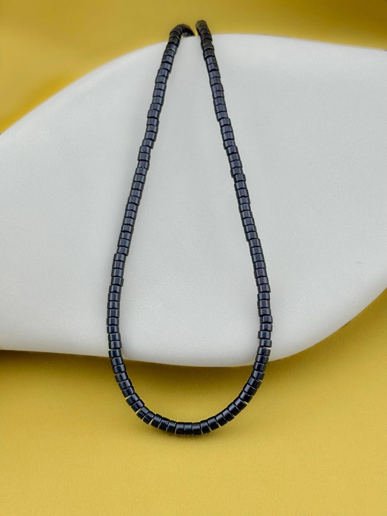 May include: A single strand necklace composed of small, cylindrical, dark charcoal-colored beads. The beads are uniform in shape and size, strung together to form a continuous loop that hangs in a simple, elongated oval shape against a smooth surface.