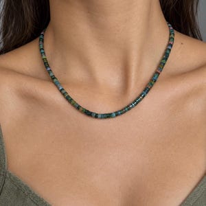May include: A beaded necklace featuring small, cylindrical, multi-colored stones in shades of deep blue, green, and earthy brown. The beads are strung together in a continuous, uniform pattern, creating a slim, circular strand that rests around the neck.