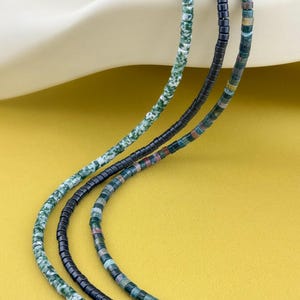 May include: Three strands of beaded necklaces featuring small cylindrical beads. One strand consists of mottled green and white beads, another of solid black beads, and the third of multicolored beads in shades of green, brown, and grey.